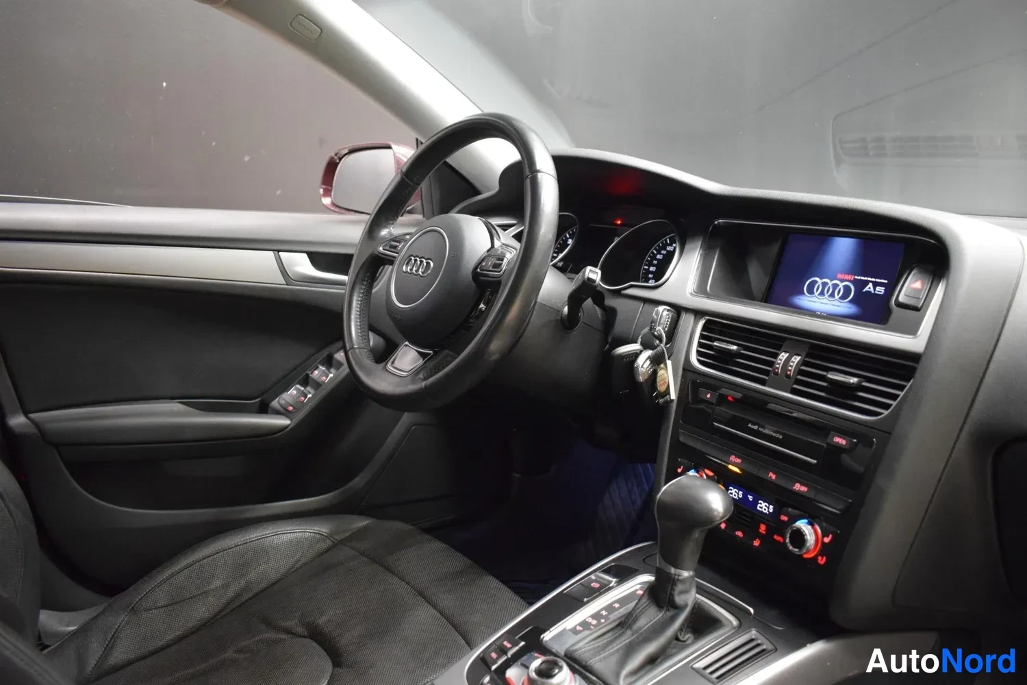 Muu: Auton etusivun kuljettajan puolen sisätila: Audi-ohjauspyörä, kojetaulu, infotainment-näyttö, automaattivaihteiston valitsin ja tumma urheiluistuin.