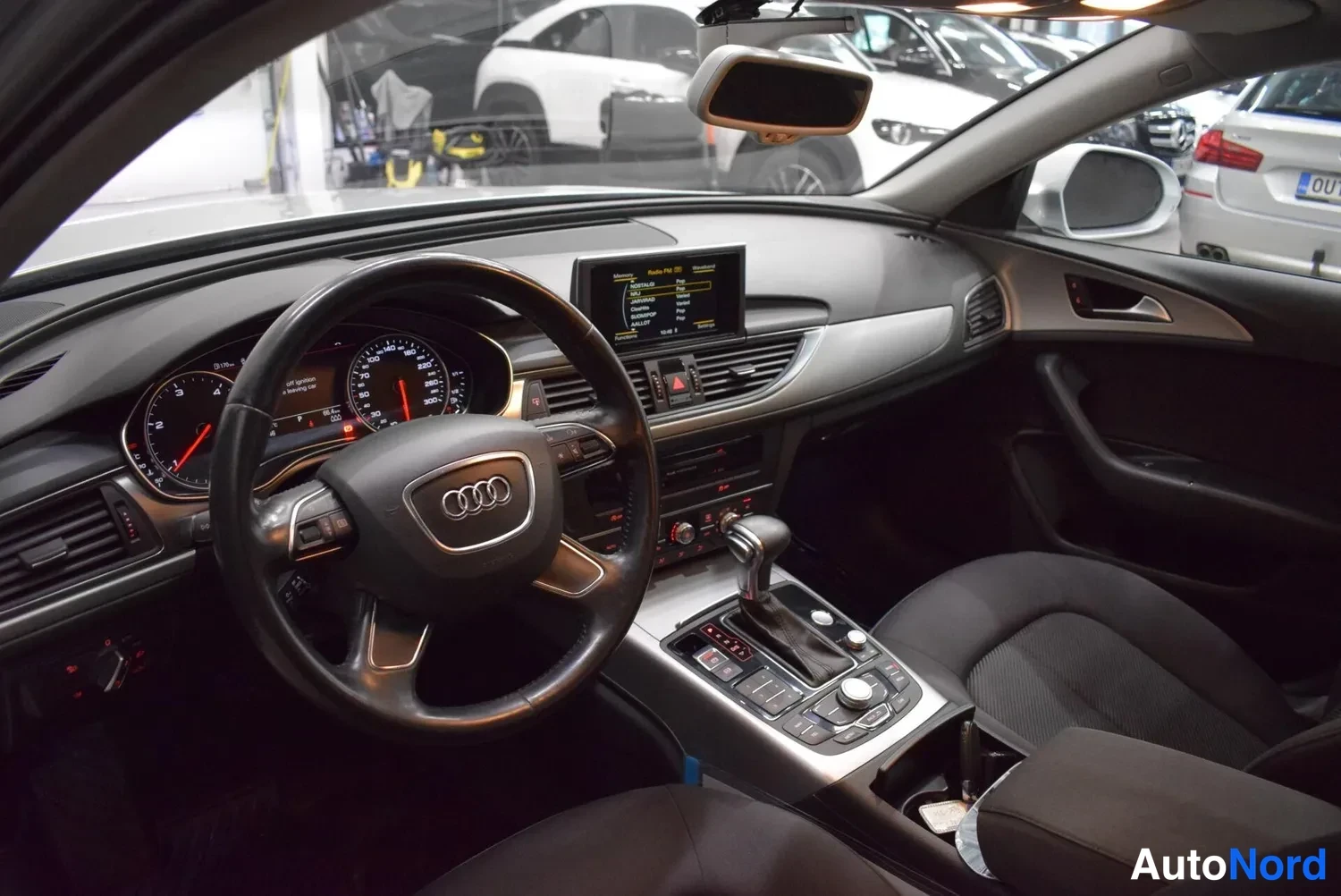 Muu: Auton sisätila ohjaamosta: Audi-ohjauspyörä, mittaristo, keskikonsolilla näyttö ja vaihteenvalitsin, mustat istuimet.
