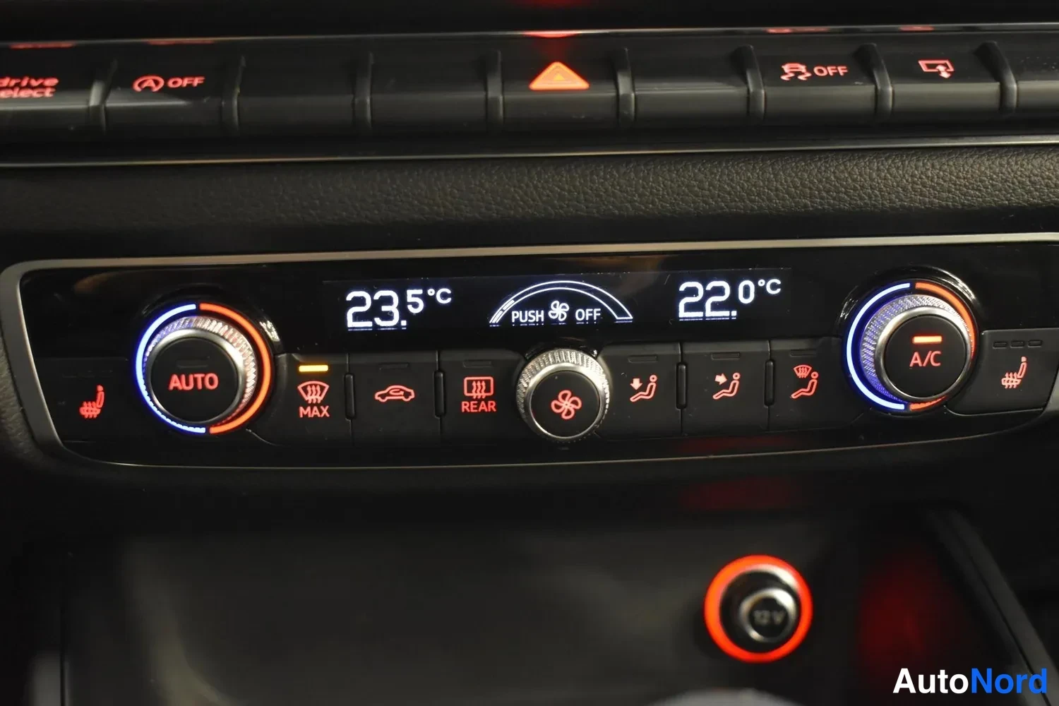 Muu: Keskikonsolin ilmastointipaneeli lähikuvassa: AUTOn ja A/C:n pyöröt, lämpötilat 23,5 °C ja 22,0 °C, MAX- ja REAR-napit sekä 12 V-pistoke.