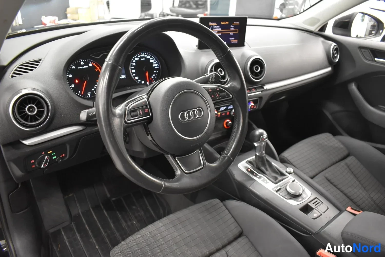 Muu: Audi-henkilöauton kuljettajan näkymä: ohjauspyörä Audi-merkillä, mittaristo, automaattivaihteen valitsin ja keskikonsolin näyttö.