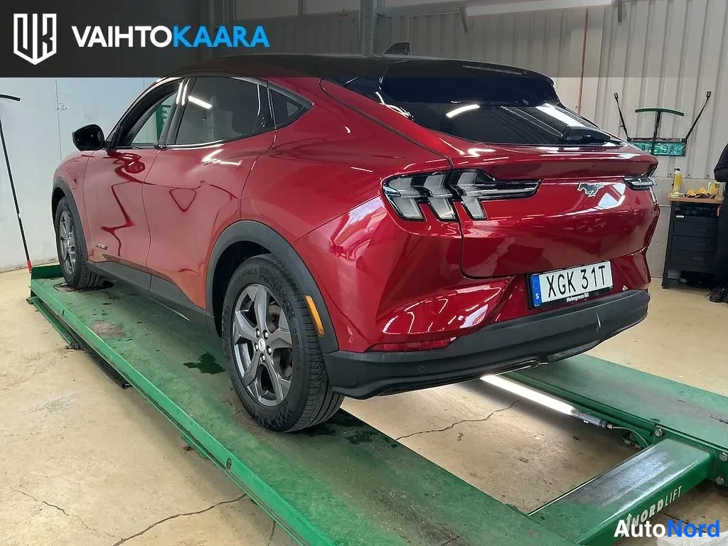 Takaviisto: Punainen Ford Mustang Mach-E takaosa kolmivälineestä oikealle kääntyneenä nosturin päällä, rekisteritunnus XGK 31T.