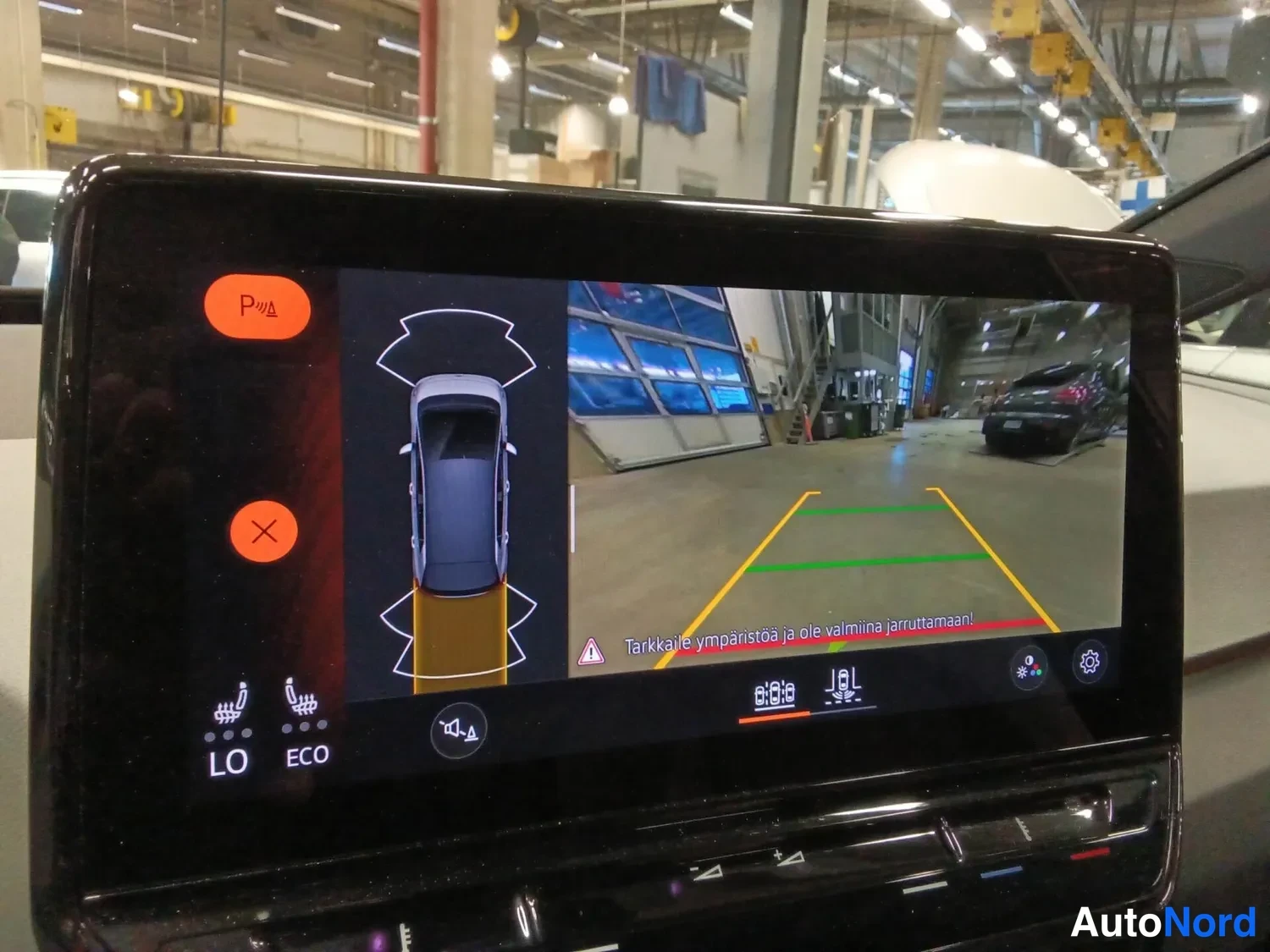 Muu: Infotainment-näyttö, jossa peruutuskameran kuva pysäköintitilanteesta ja pysäköintianturien grafiikka. Näytössä oranssi P-painike ja varoitusteksti.