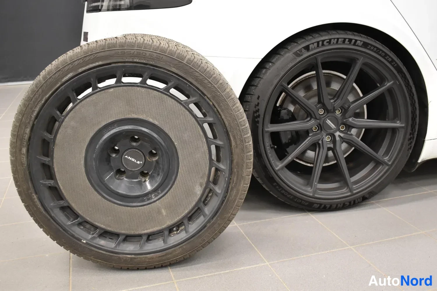 Muu: Renkaat: Michelin talvirengas 285/30R21 (vasemmalla) ja musta vanteella varustettu rengas (oikealla), laatoitettu lattia taustalla.