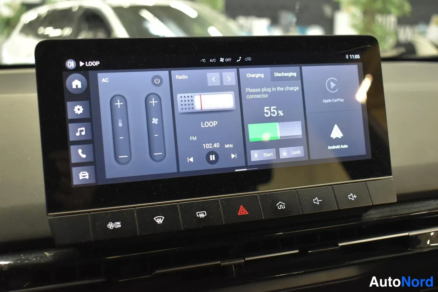 Yksityiskohta: Keskikonsolin kosketusnäyttö ilmastointi-, radio- ja lataustietojen kanssa; lataus 55 %, radiokanava FM 102.40 MHz. Apple CarPlay- ja Android Auto-ruudut näkyvä
