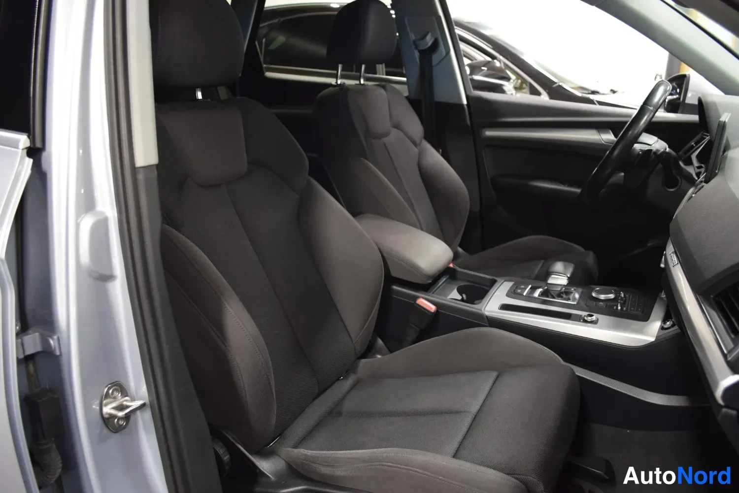 Edestä: Audi Q5:n etusisätilat kuvattuna avatusta etuovesta: mustat kangasistuimet, keskikonsolin vaihdekeksi ja infotainment-näyttö sekä ohjauspyörä.