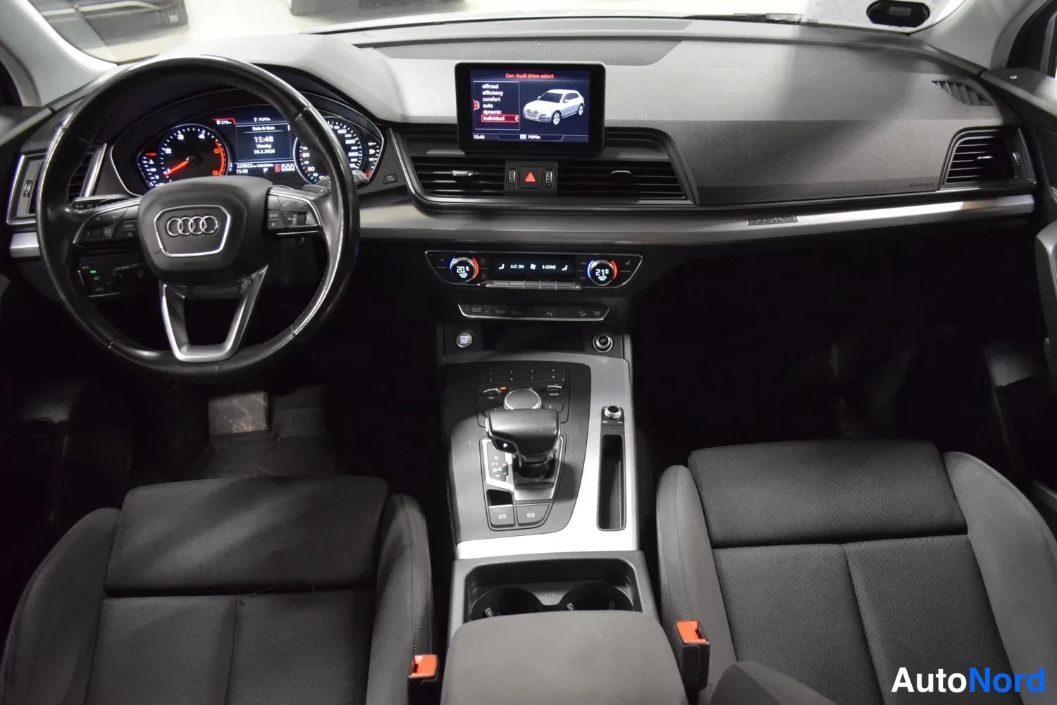 Sivulta: Audi Q5:n ohjaamo kuvattuna etuistuinten välistä: Audi-ohjauspyörä,mittaristo, keskikonsolin automaattivaihteen valitsin ja päällä oleva infotainment-näyttö, jo