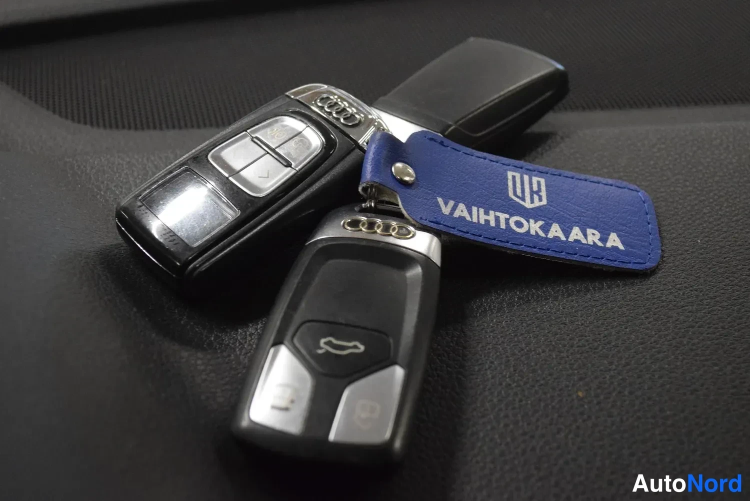 Yksityiskohta: Kaksi Audi-avain.nippua tummalla pinnalla, mukana sininen VAIHTOKAARA-avain.nippu.