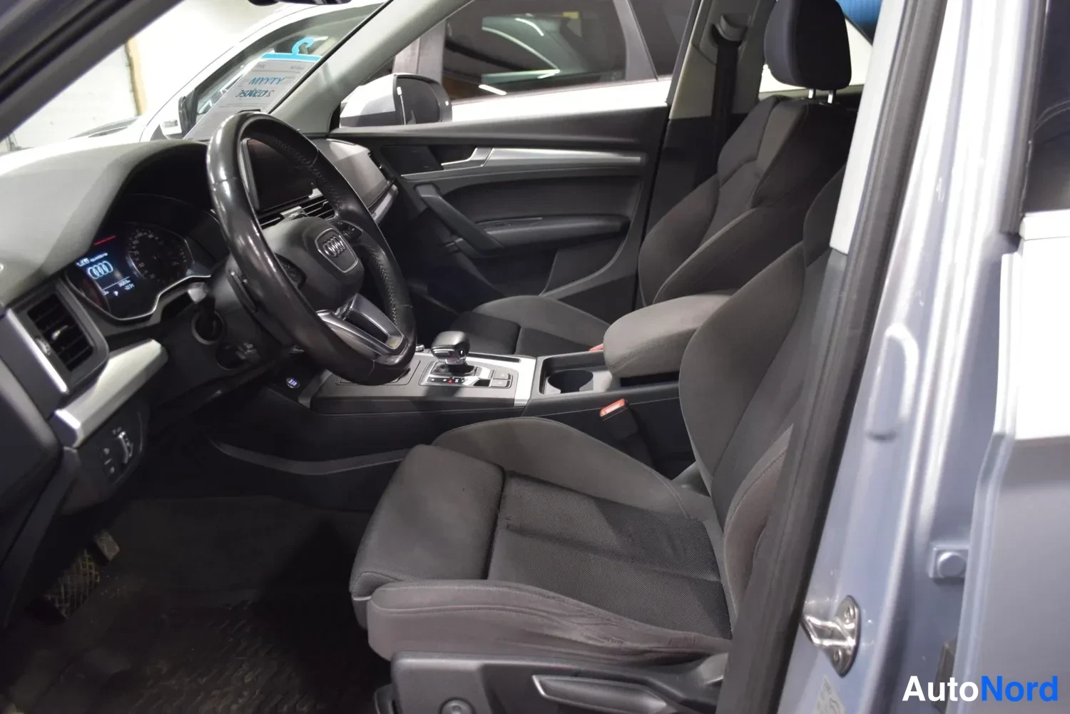 Sivulta: Audi Q5:n sisätilakuva kuljettajan ovesta: tumma kankaaverhoilu, ohjauspyörä ja kojelauta, viisiovinen matkustamomiljöö.