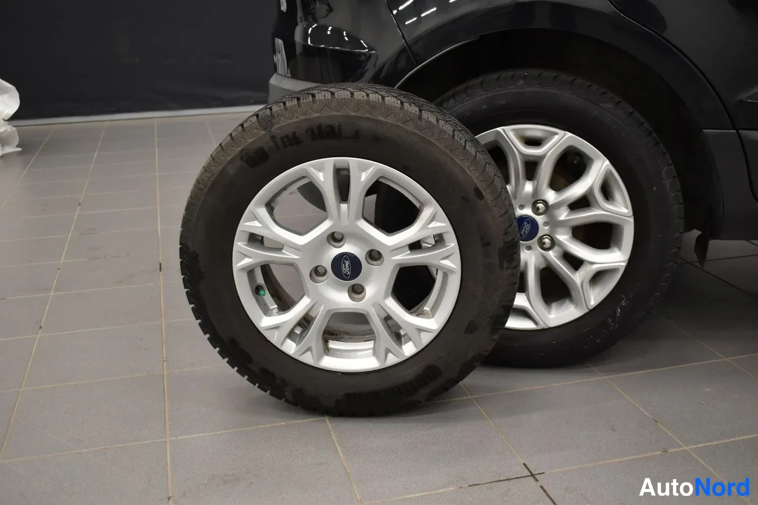 Yksityiskohta: Kaksi rengasta hopeavanteella: etualalla irtonainen rengas ja auton takana kiinnitetty rengas, Ford-merkki napassa.