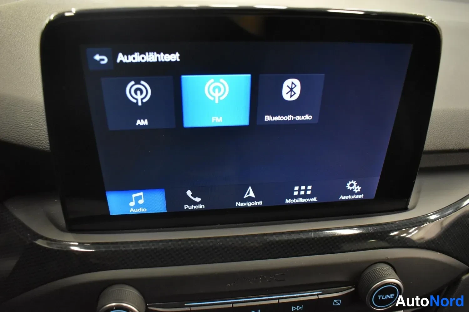 Muu: Fordin infotainment-näyttö Aviolähteet-valikossa, AM/FM ja Bluetooth-audio vaihtoehtoina; alapalkissa Puhelin, Navigointi ja Asetukset.