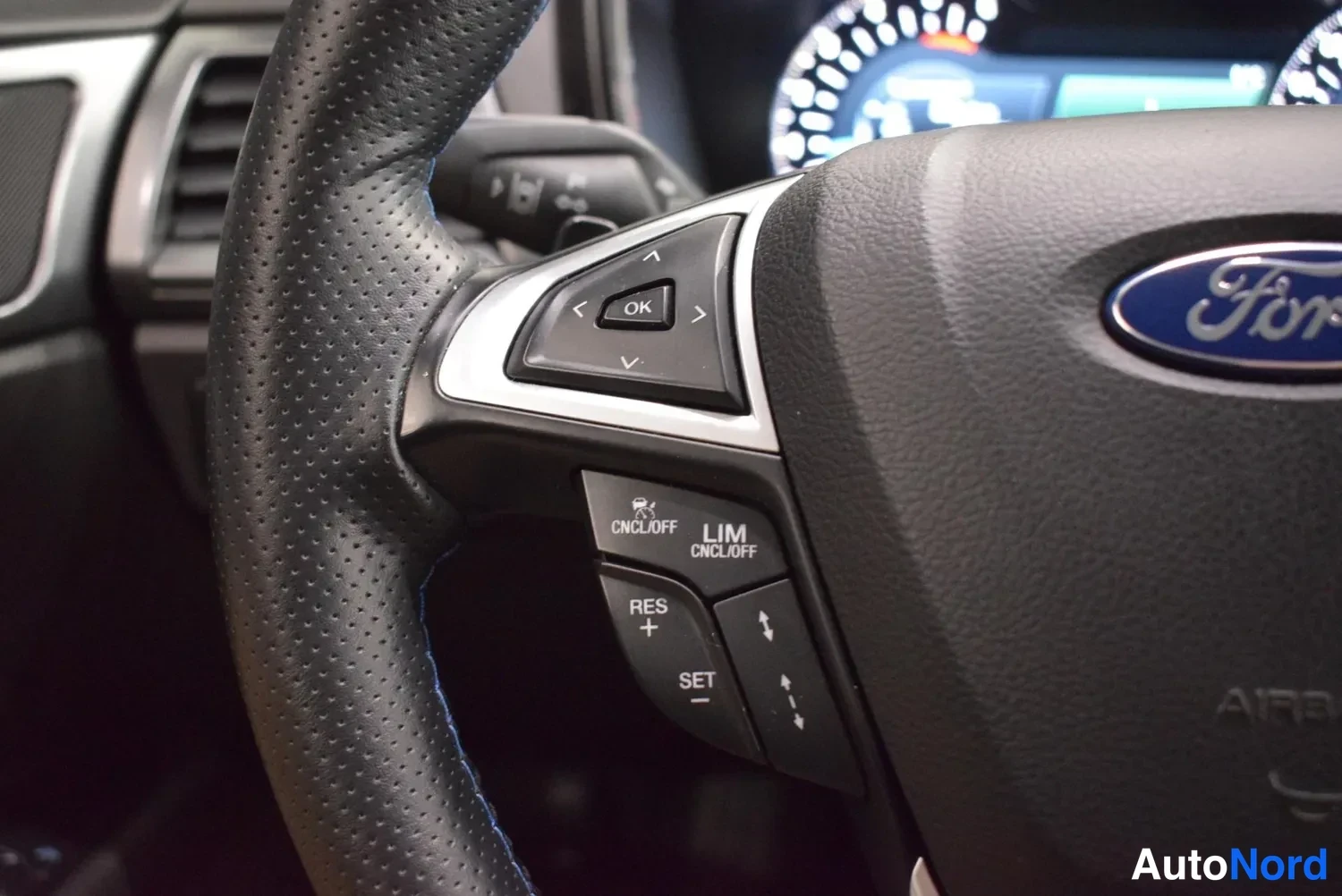 Edestä: Ford-ohjauspyörä lähikuva: merkkilogonmuste keskellä ja monitoiminäppäimet CRUISE cncl/off, lim/lim/off, res+, set- ja nuolinäppäimet.