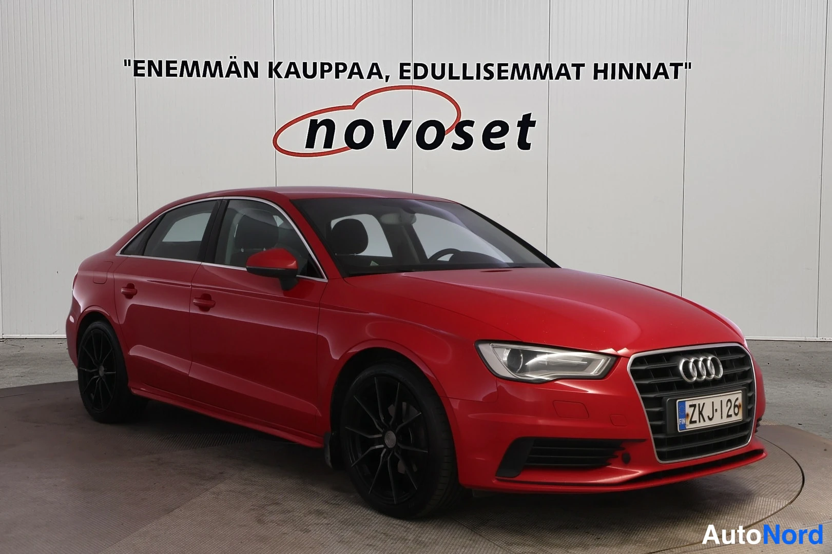 Etuviisto, nokka oikealle: Punainen Audi-sedan kolmessa neljäsosassa edestä oikealta, mustat kevytmetallivanteet, rekisterikilpi.