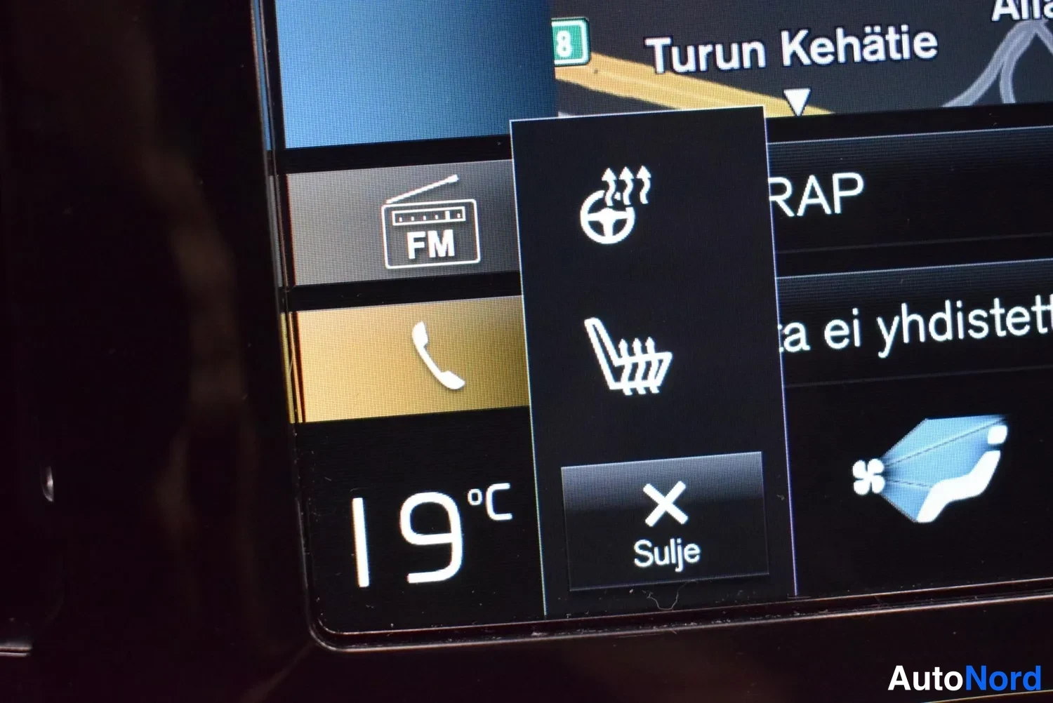 Yksityiskohta: Lähikuva infotainment-näytöstä: ponnahdusvalikko ohjaustatten ja istuinten lämmityskuvakkeista, 'Sulje'-painike, FM-radio ja lämpötila 19 °C.