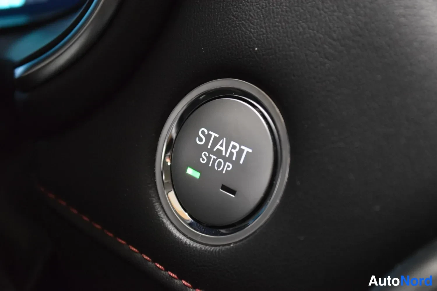 Yksityiskohta: Start/stop-nappi tummassa kojelaudassa, vihreä merkkivalo symboleineen.