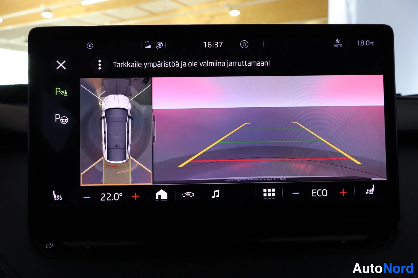 Yksityiskohta: Infotainment-näyttö näyttää 360°-kameran ylhäältäkatsekuvan autosta ja peruutuskameran opastusviivat; ruudulla varoitus 'Tarkkaile ympäristöä ja ole valmiina ja
