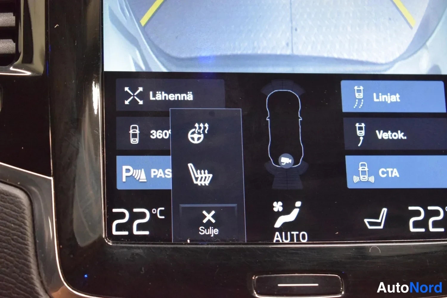 Muu: Yksityiskohta auton infotainment-näytöstä: takakameran kuva ja kosketusvalikko, jossa vaihtoehdot Lähennä, 360°, PAS ja Linjat, Vetok., CTA sekä lämpötilat 22 °