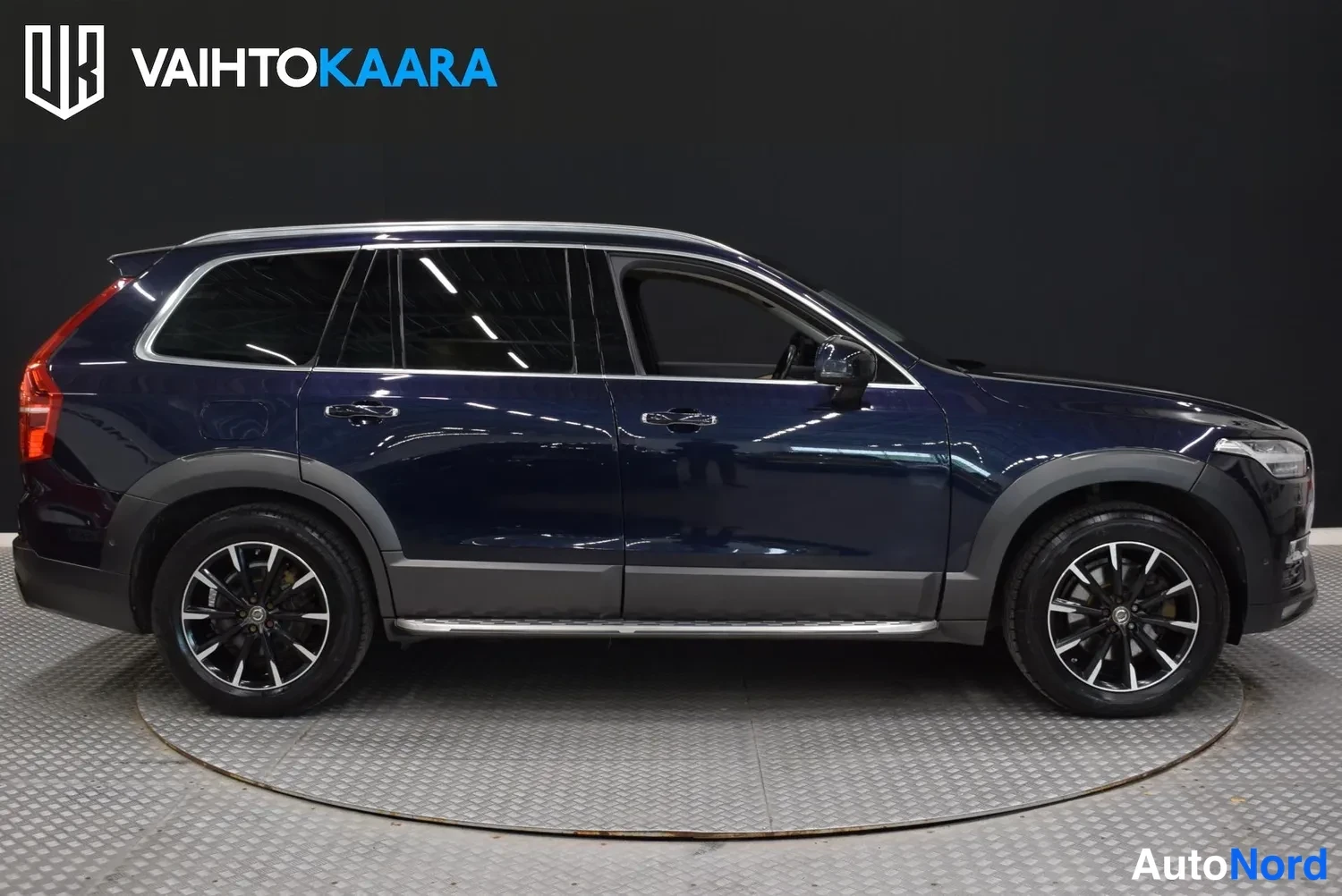 Etuviisto, nokka oikealle: Tummansininen Volvo XC90 -maasturin sivunäkymä, oikea kylki ja keula kuvassa, aitoissa pyörissä.