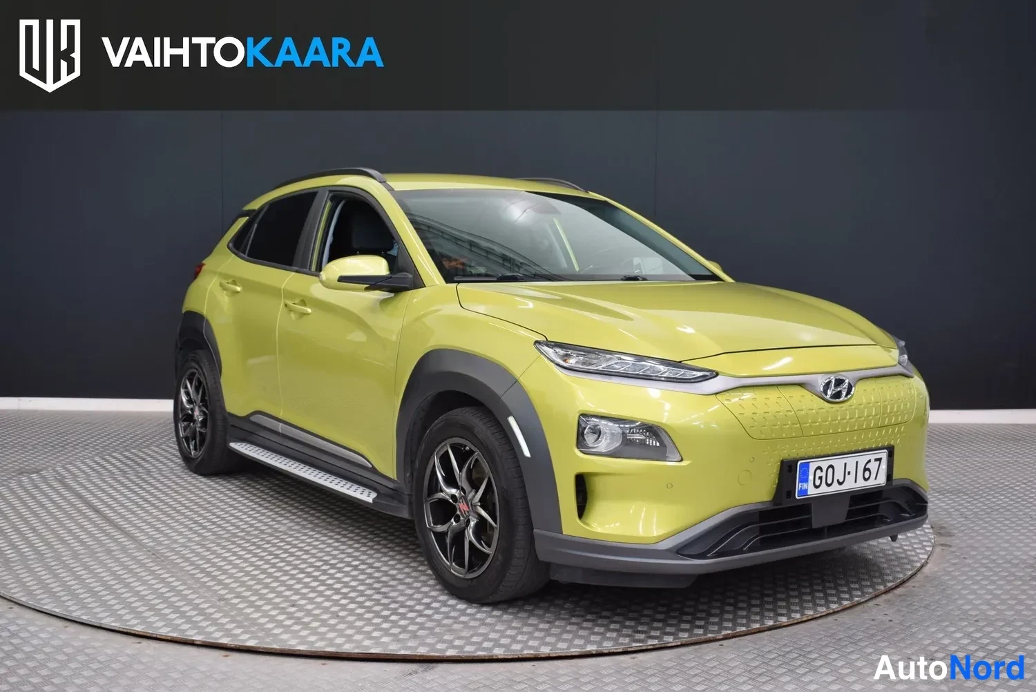 Etuviisto, nokka oikealle: Hyundai Kona Electric etukulmasta oikealta, keltainen kori, rekisterikilpi, LED-ajovalot ja mustat kevytmetallivanteet.