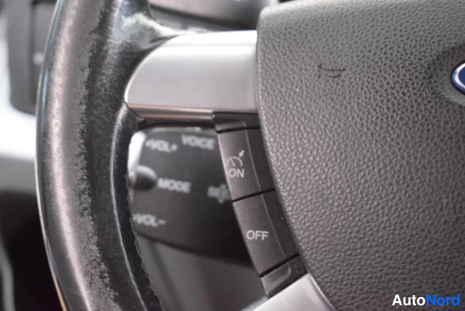 Muu: Ohjauspyörän vasen puoli lähikuvassa, jossa ratissa olevat painikkeet: VOLUME+, VOICE, MODE, VOLUME-, ja erillinen kytkin ON/OFF (hätäkilvet). Ford-logon reuna