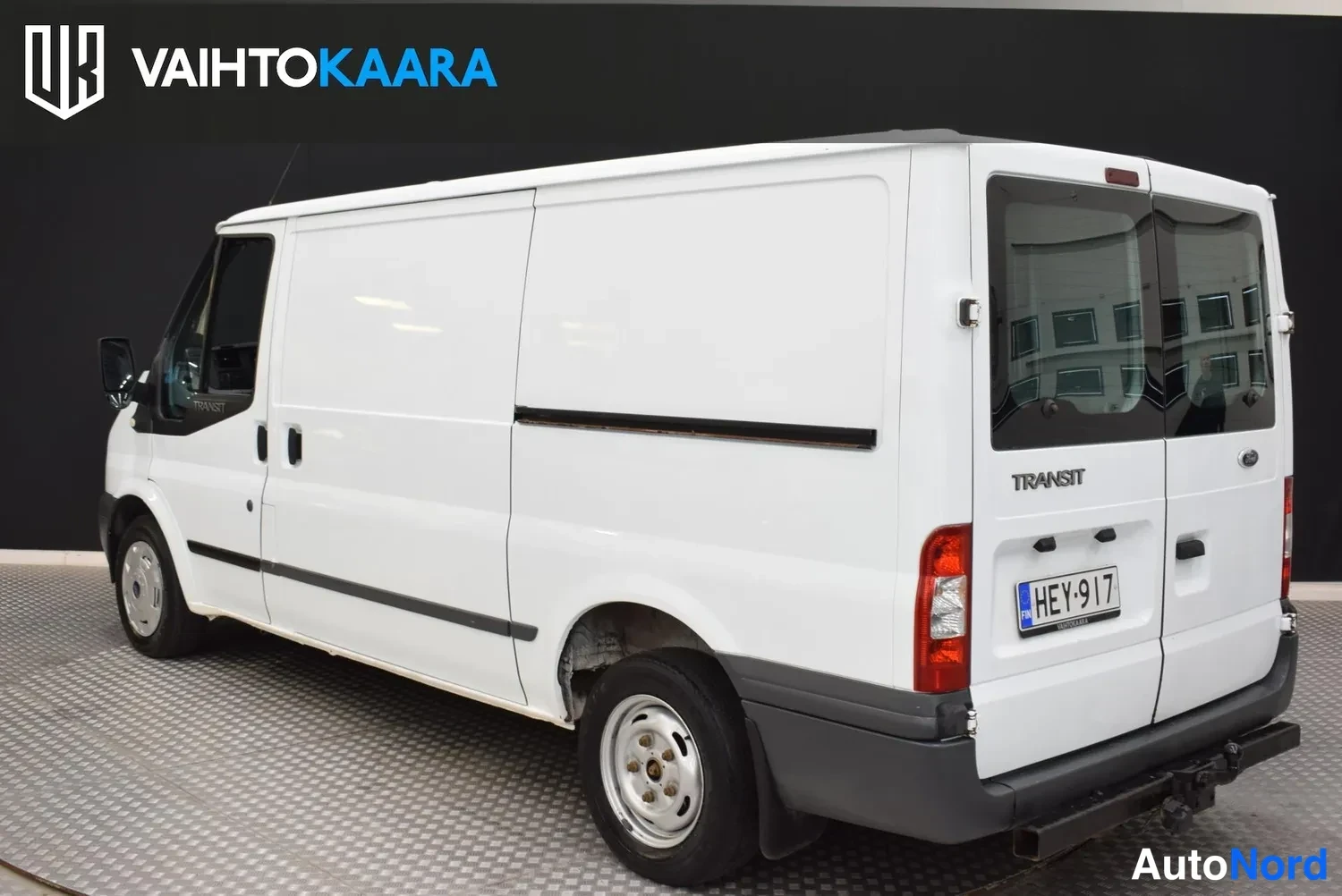 Takaviisto: Ford Transit -pakettiauto valkoisena takaosan kulmasta, Transit-teksti ja rekisterikilpi.