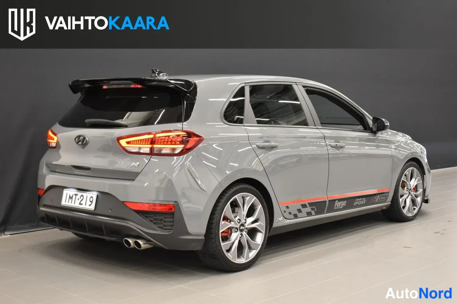 Takaviisto: Hyundai i30 -viistopyykkö harmaana takanäkymä toiselta sivulta, musta spoileri, kaksi pakoputkea ja punaiset takavalot näkyvät.