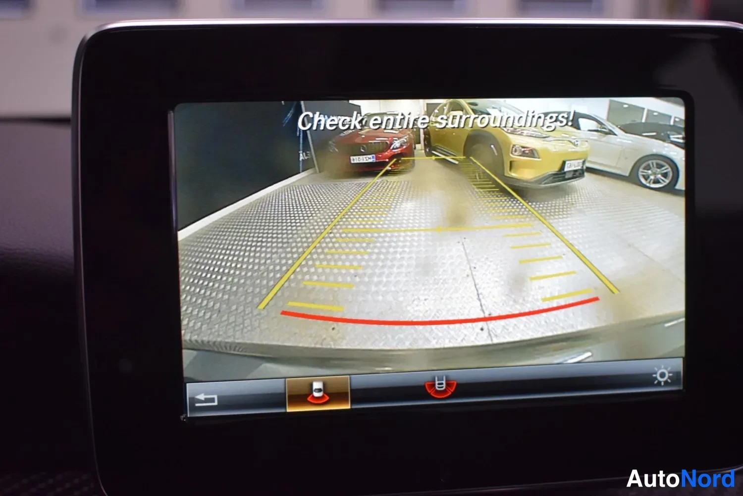 Muu: Infotainmetscreenin käänteinen peruutuskameranäkymä, jossa keltainen ja punainen parkkipaikkaohjain ja teksti 'Check entire surroundings!'.