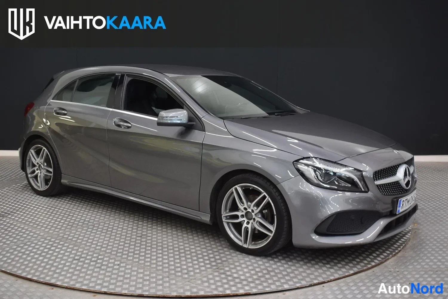 Etuviisto, nokka oikealle: Hopeanharmaa Mercedes-Benz A -luokan viistosti edestä oikealta kuvattuna hallissa pyörivällä tasolla, AMG-tyyliset keulakotelo ja viisisakarainen kevytme