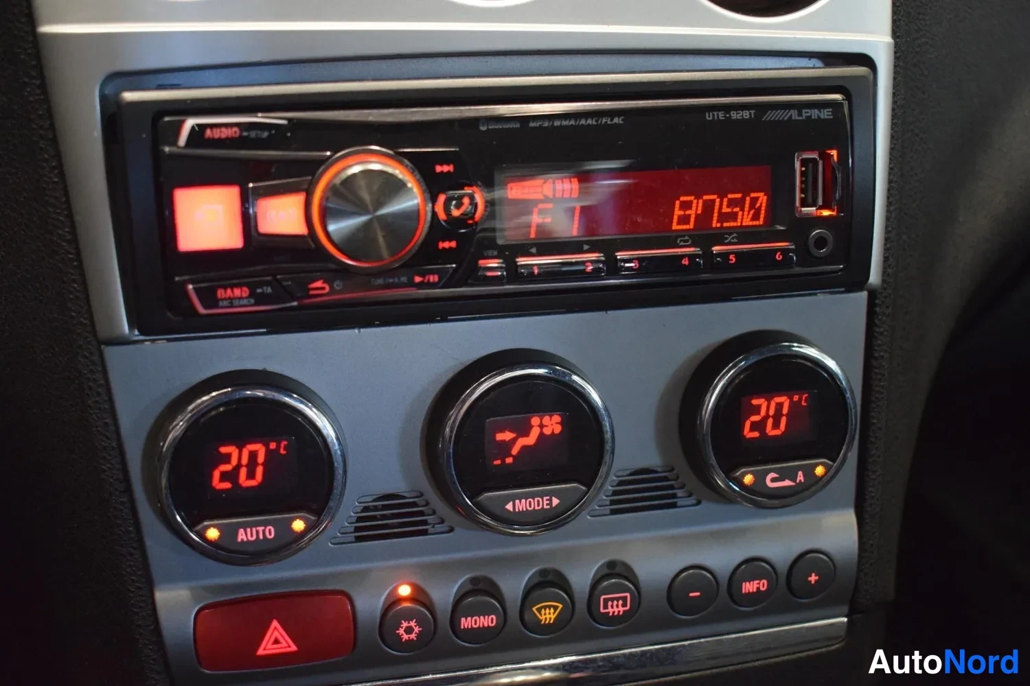 Yksityiskohta: Keskikonsolin lähikuva: Alpine -radiosoitin punaisella näytöllä (arvo 87.50), alla kolme pyöreää ilmastoinnin säädintä, joissa lukema 20°C ja AUTO- sekä MODE-te