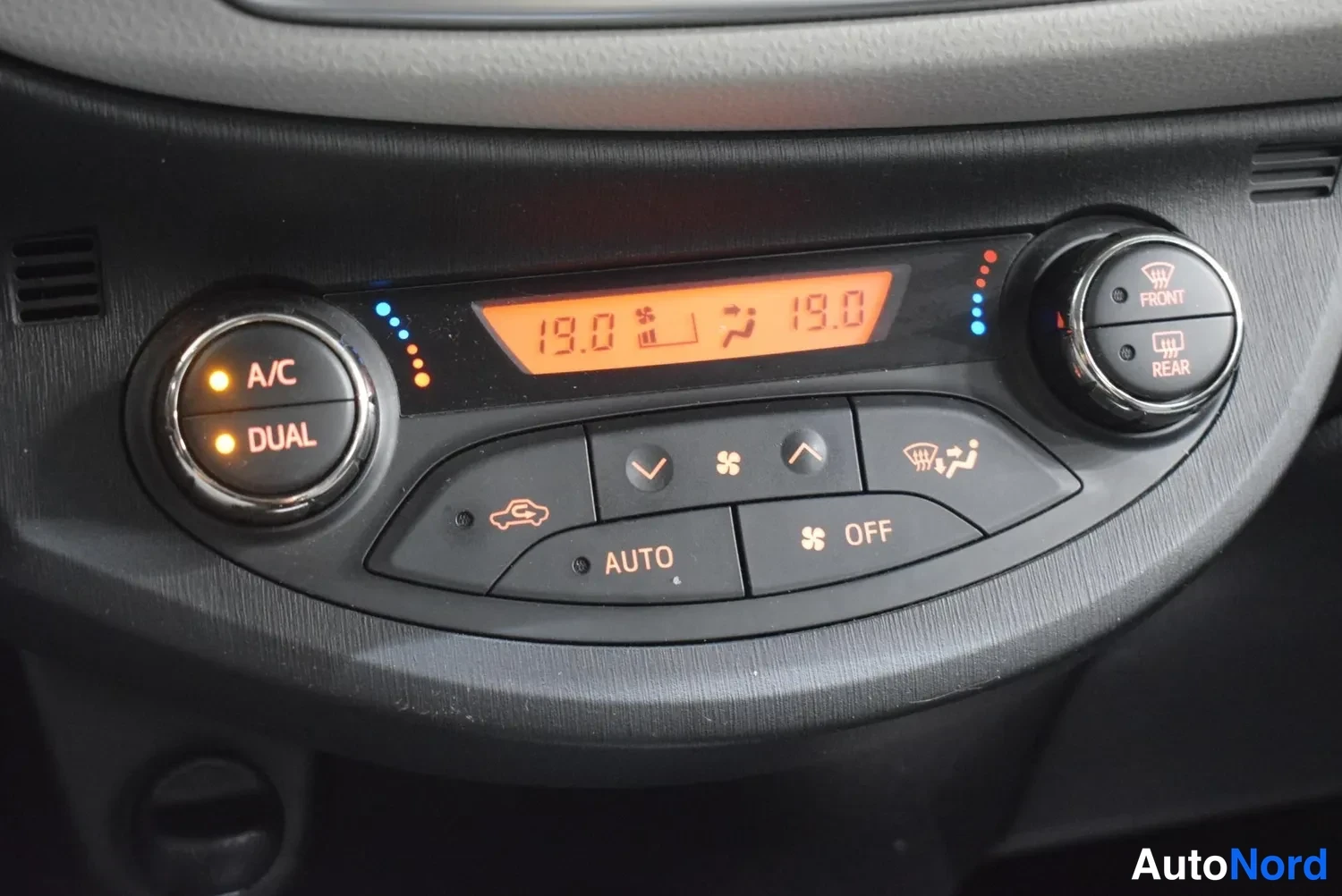 Muu: Ilmastointipaneeli lähikuvassa: A/C ja DUAL-painikkeet valaistuina, näyttö 19.0 astetta molemmin puolin, AUTO- ja OFF-painikkeet, etu- ja takaistuimen lämmitys.