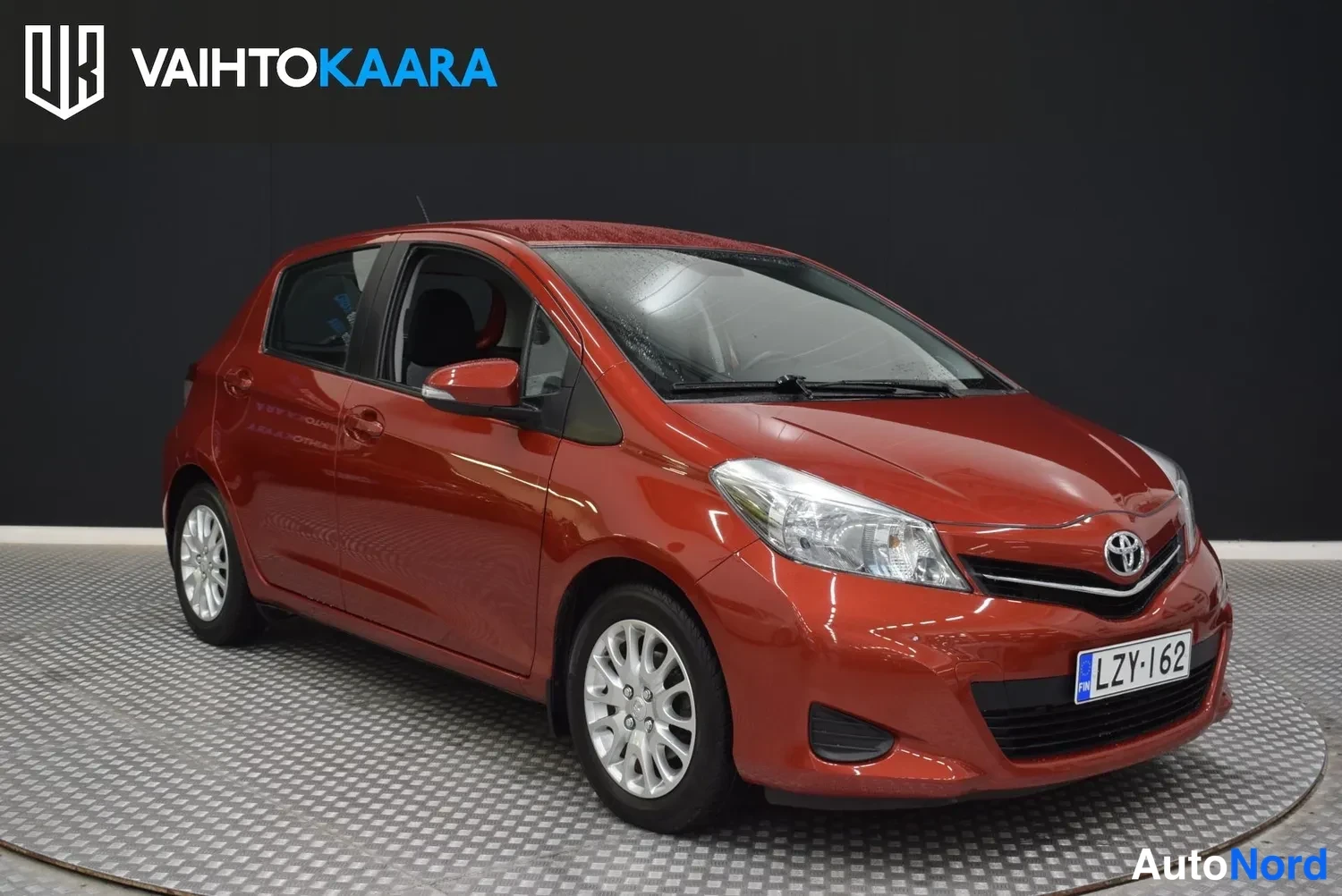 Etuviisto, nokka oikealle: Punaisen Toyota Yaris -henkilöauton etukulma oikealta, etupyörät ja rekisterikilpi näkyvissä.