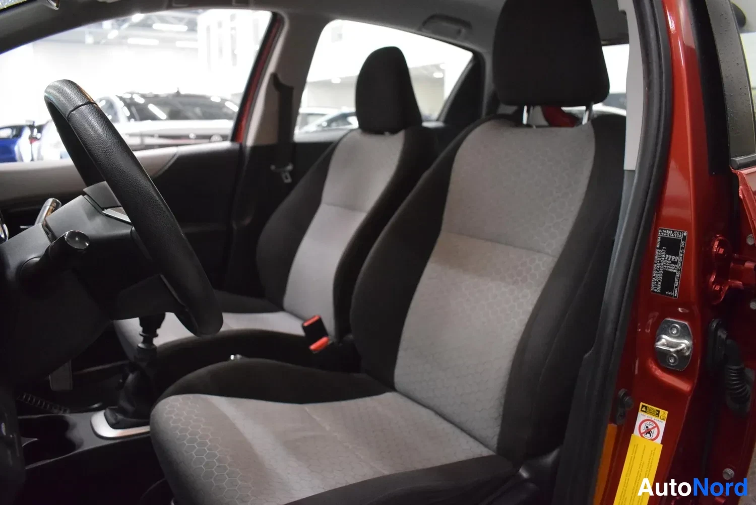 Muu: Sisäkuva Toyota Yaris -henkilöauton etusohvasta: kaksi harmaa-mustaa kankaista etuistuinta, ohjauspyörä ja vaihtehyksy, punainen kori näkyy avatusta ovenkannest
