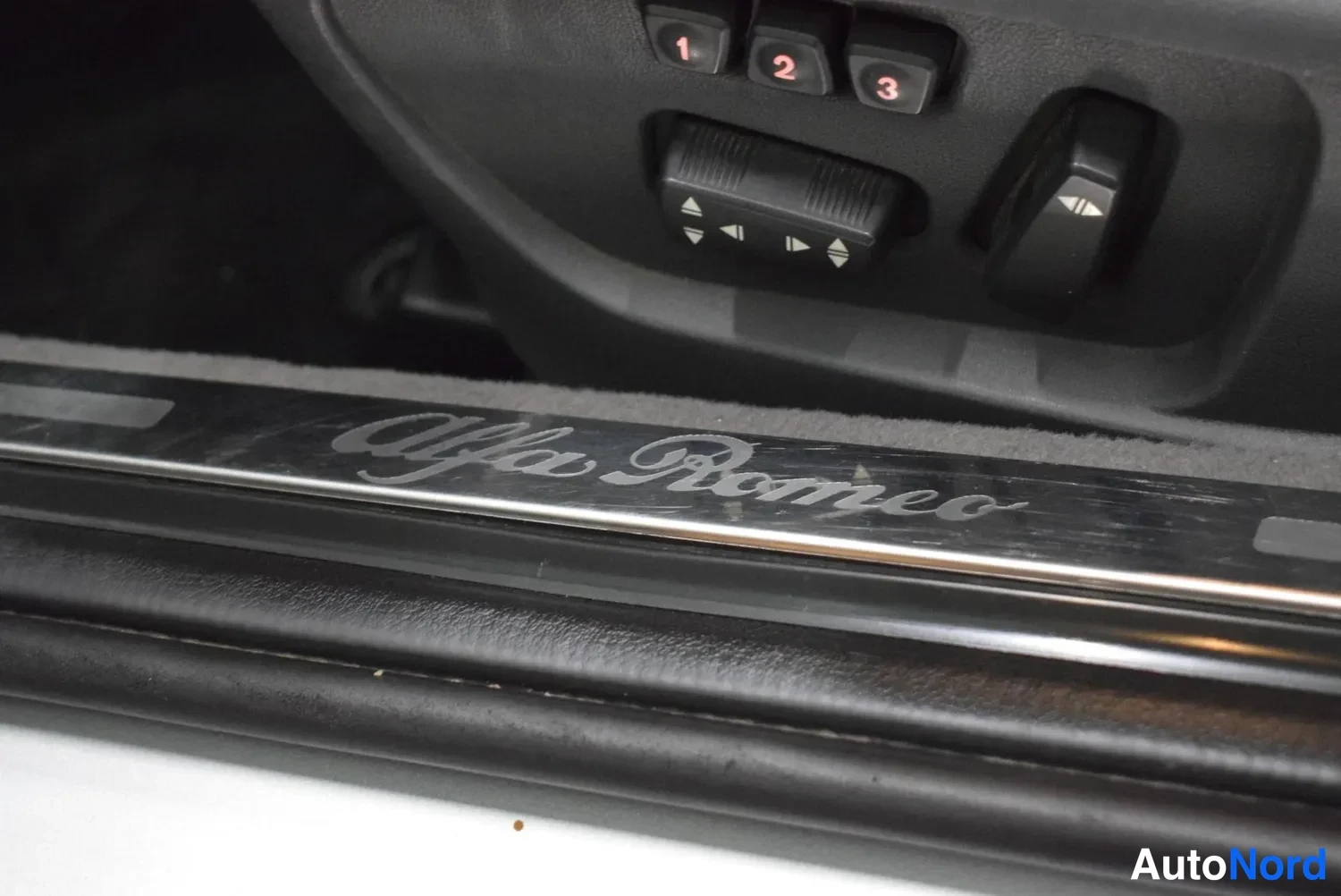 Yksityiskohta: Auton ovikynnyslistassa teksti Alfa Romeo, ylhäällä istuimen muistipainikkeet 1, 2 ja 3 sekä peilin säätökytkimet.
