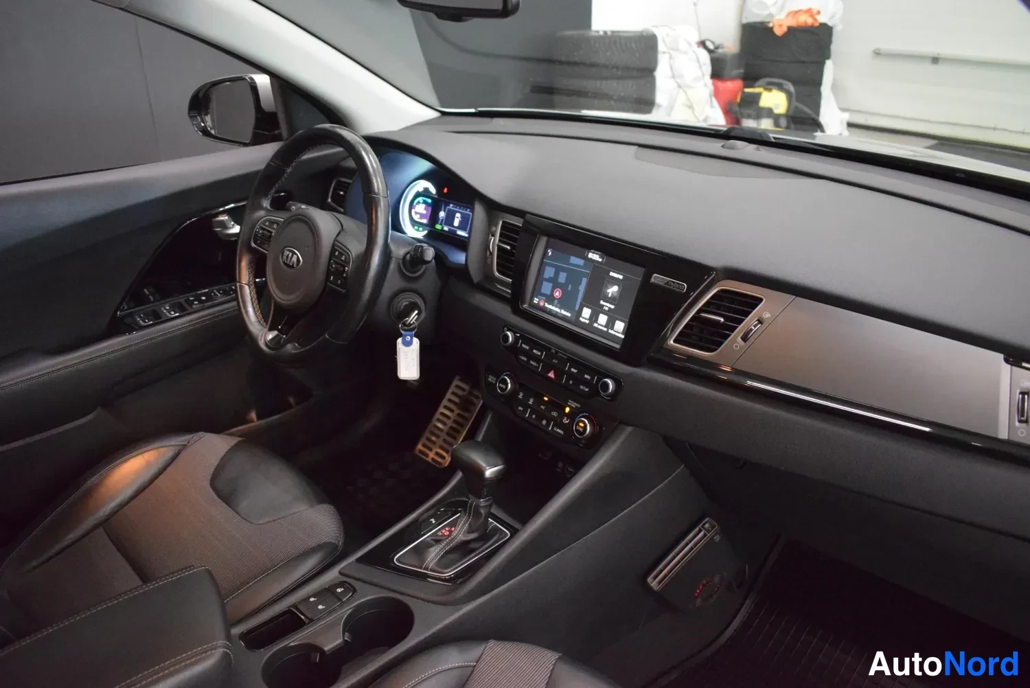 Edestä: Auton sisätilat: ohjaamo, jossa Kia-merkkinen ohjauspyörä, infotainment-näyttö, automaattivaihteen valitsin ja mittariston hybridinäyttö näkyvät.