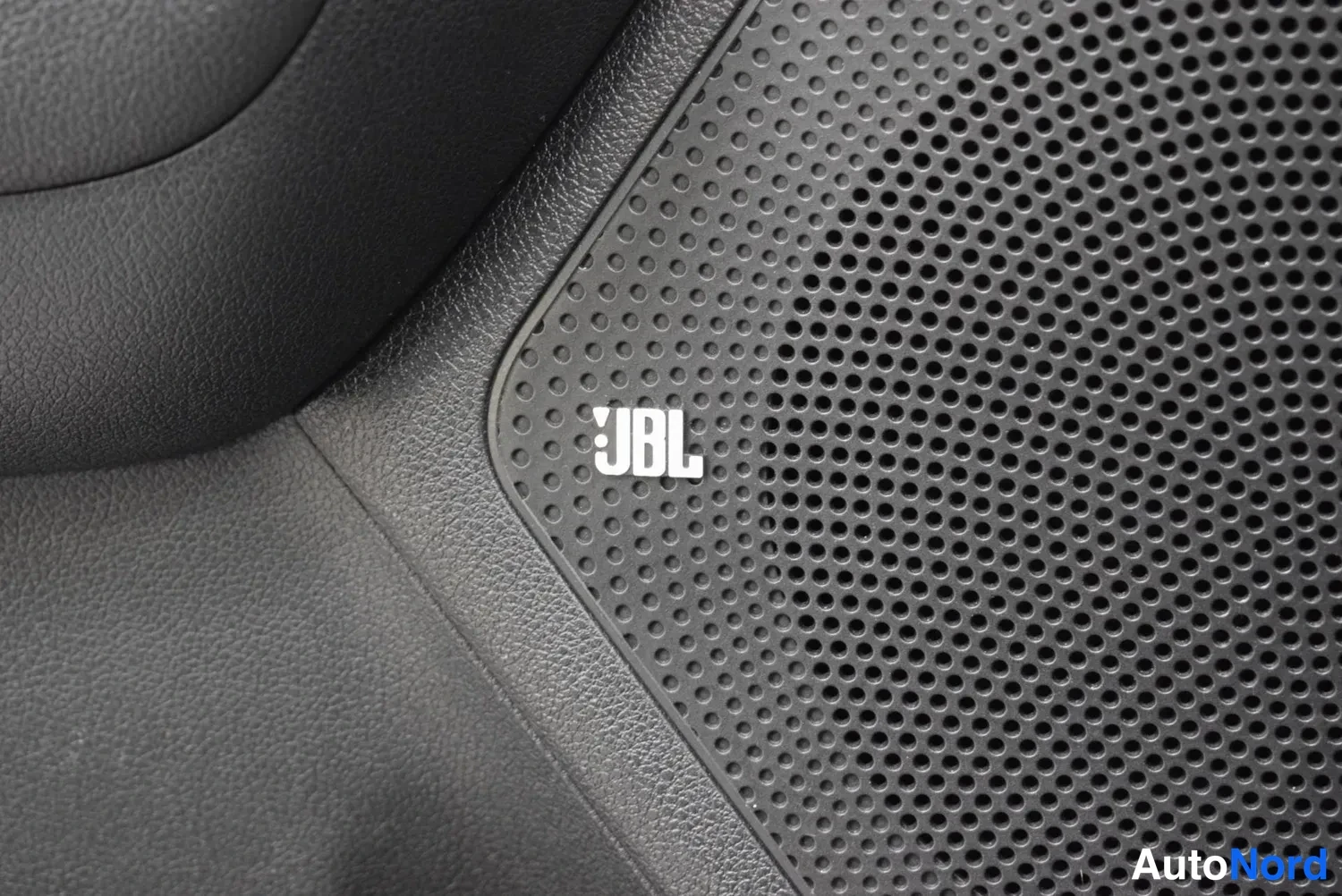 Yksityiskohta: Mustan ovenpaneelin kaiuttimen verkko lähikuvassa; siinä on valkoinen JBL-logo.