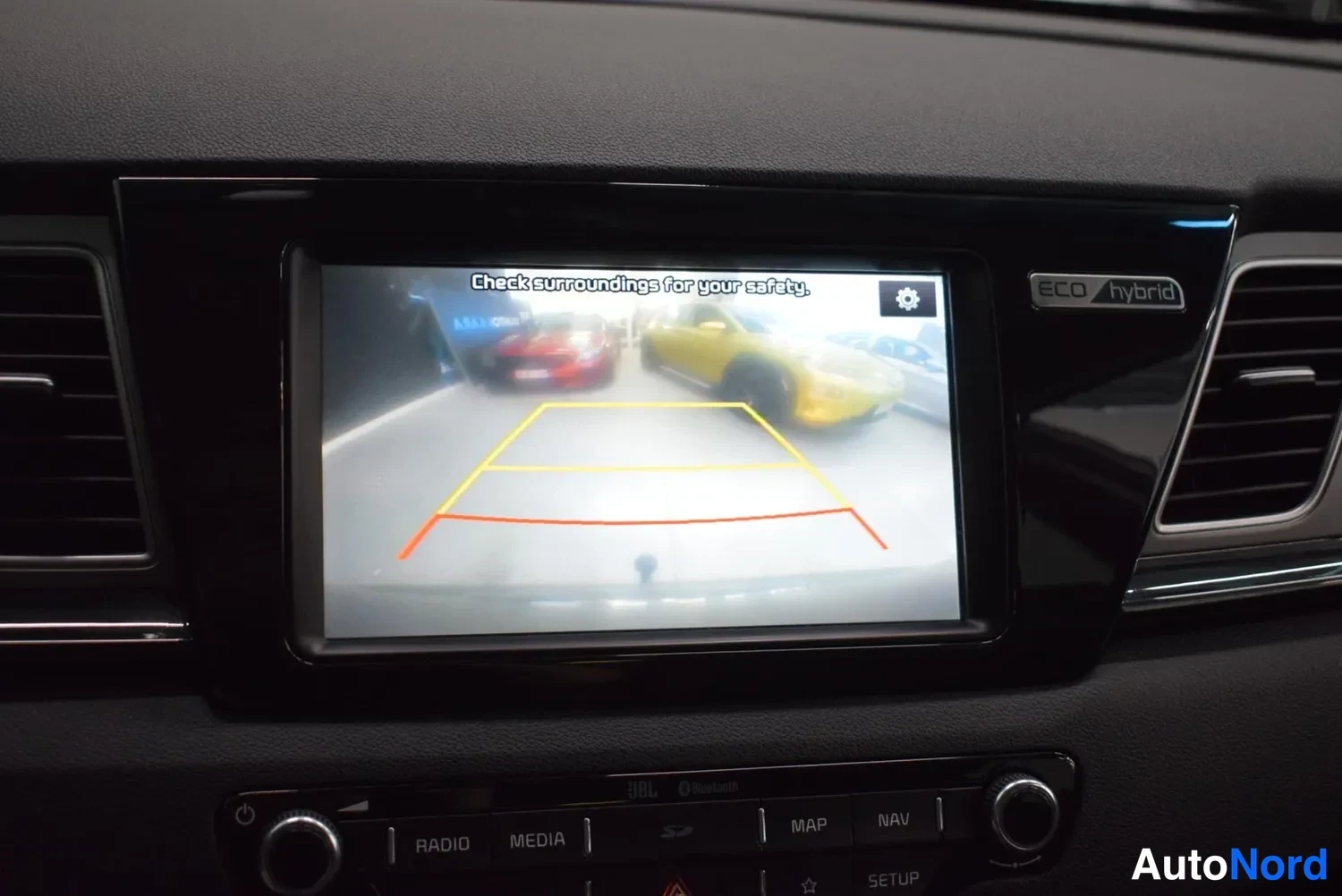 Yksityiskohta: Keskikonsolin näytöllä näkyy peruutuskamera: kuvassa varastohallia autoineen ja oranssit/keltaiset etäisyysviivat, otsikolla 'Check surroundings for your safety