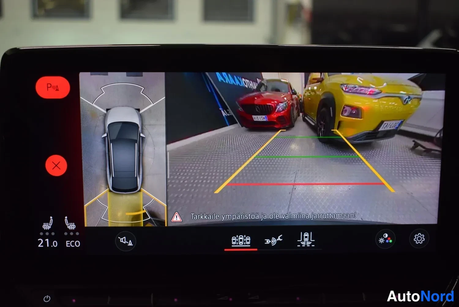 Yksityiskohta: Auton infotainment-näyttö näyttää 360-kaapelinäkymän peruutettaessa, teksti Tarkkaile ympäristöä ja ole valmiina jarruttamaan!