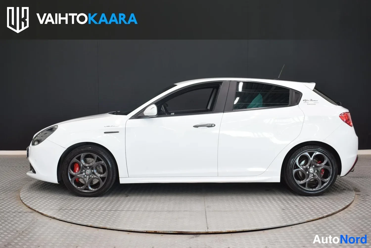 Sivulta: Valkoisen Alfa Romeo Giulietta -henkilöauton sivuprofiili studiotummaa taustaa vasten, tummat kiinteät vanteet ja punaiset jarrusatulat näkyvät.