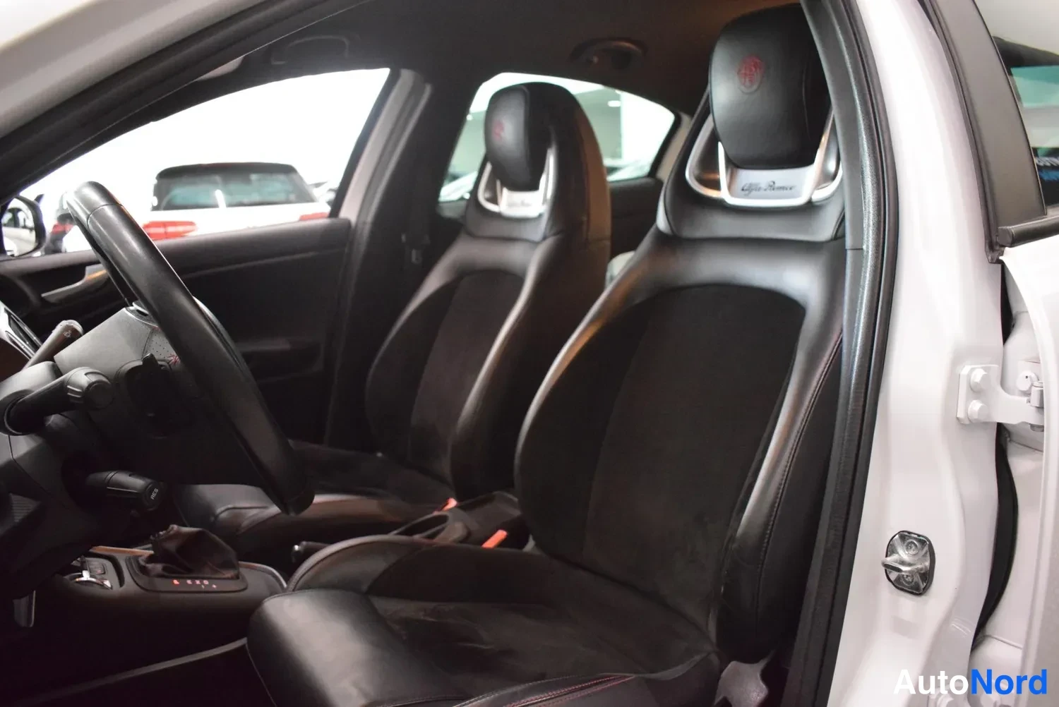 Edestä: Auton sisätila: kaksi mustaa urheiluistuinta, joissa Alfa Romeo -logot alustuissa, ohjauspyörä ja avattu sivuovi näkyvät vasemmalta kuvattuna.