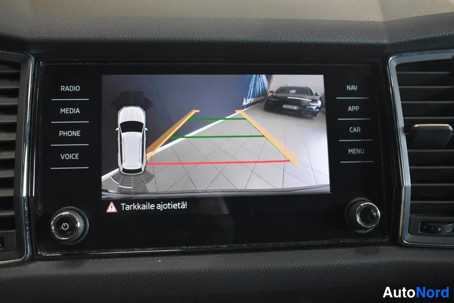 Muu: Infotainment-näyttö kääntyneenä taaksepäin; näytöllä peruutuskameran kuva pysäköintilinjoilla ja teksti 'Tarkkaile ajotietä!'.