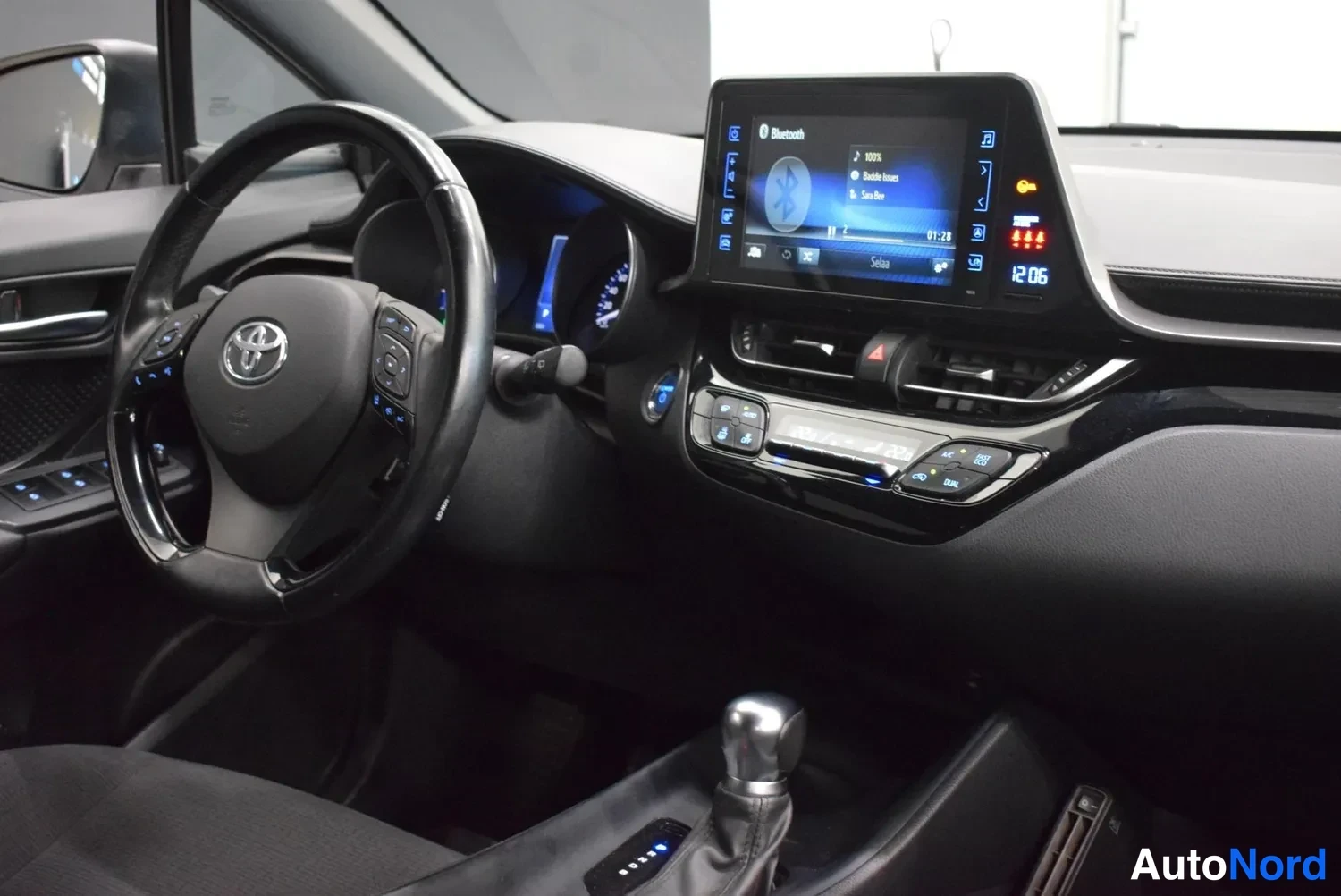 Muu: Lähikuva ohjaamosta: Toyota-ohjauspyörä, digitaalinen mittaristo ja kosketusnäyttö Bluetooth-ikkunalla, automaattivaihteen valitsin alareunassa.
