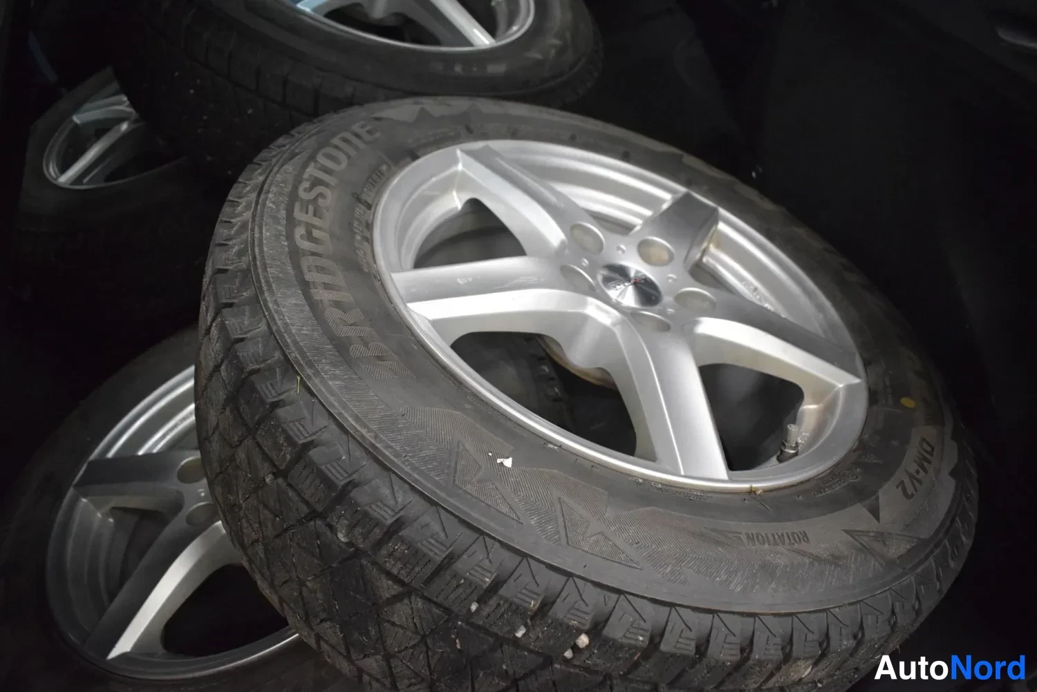 Yksityiskohta: Renkaita vanteilla pinossa: Bridgestone DM-V2 -talvirenkaan profiili ja viisipoikkeinen hopeavanne näkyvät selvästi.