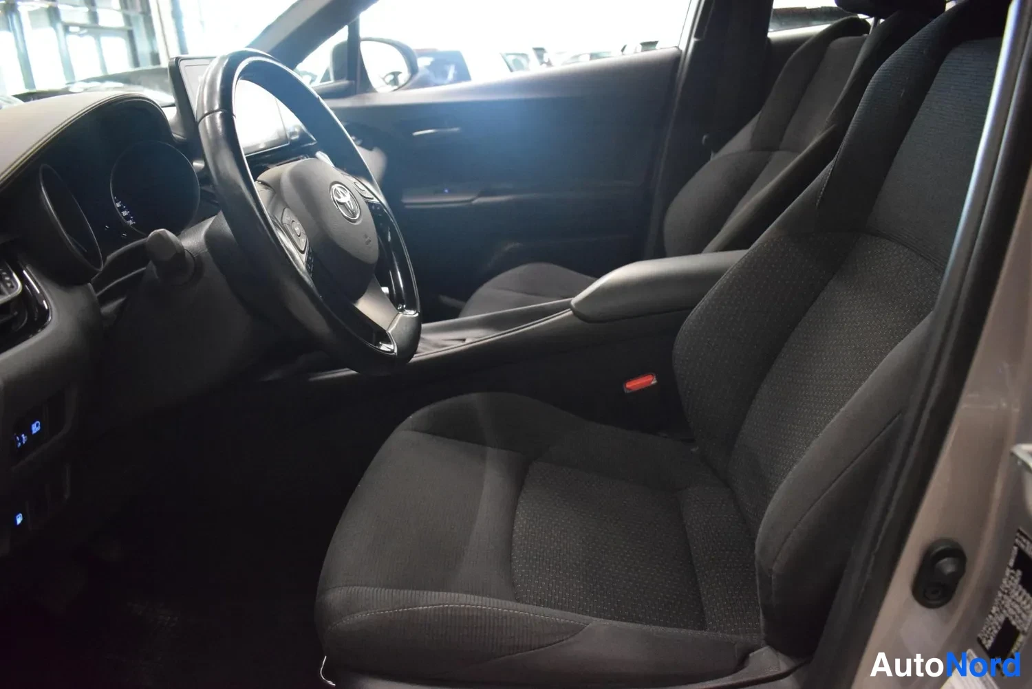 Muu: Toyota C-HR:n ohjaamo nähtynä matkustajan puolelta: tumma tekstiiliverhoilu, Toyota-merkillinen ohjauspyörä ja keskikonsolin nokka.