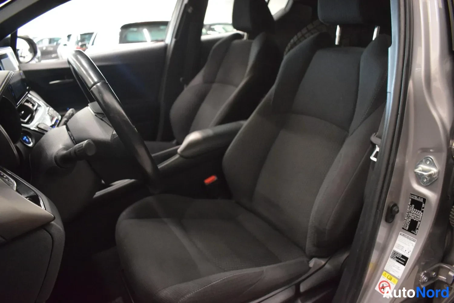 Muu: Toyota C-HR:n etuistuimet tekstiiliverhoilulla ja osa ohjaamosta, B-pilarin tyyppikilpi ja varoitustarrau näkyvät ovipuolella.