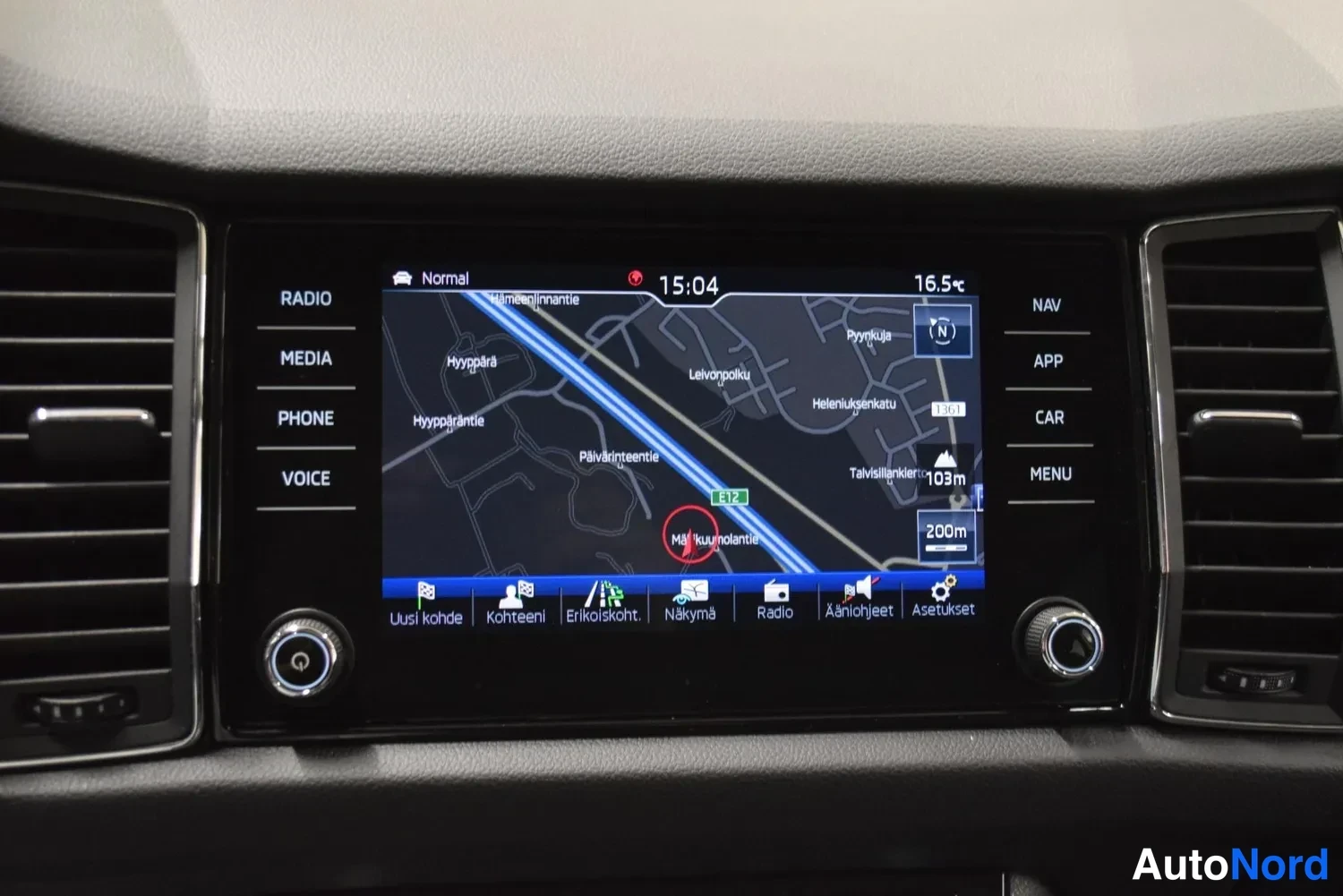 Muu: Tallenne: auton kojelaudan infotainment-näyttö näyttää navigointikarttaa ja siinä lukee Radio, Media, Phone, Voice, Nav, App, Car, Menu.