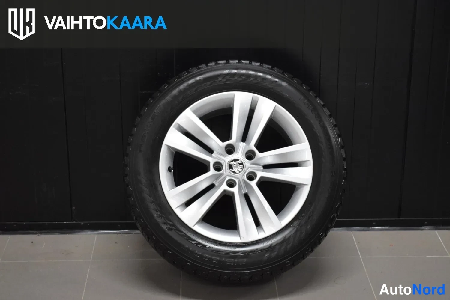 Yksityiskohta: Hopea alumiinivanne, jossa musta kitkarenkaita, mustaa seinää vasten; Skoda-logoa keskellä ja VAIHTOKAARA-logoa ylhäällä.