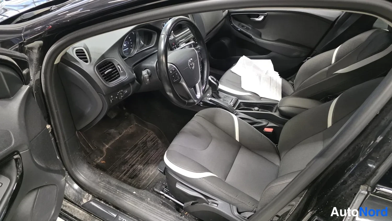 Muu: Volvo V40:n ohjaamo etupenkiltä: tummat istuimet, tekstiilityypit, jalkatilissa likaa ja paperi kuljettajan istuimella.