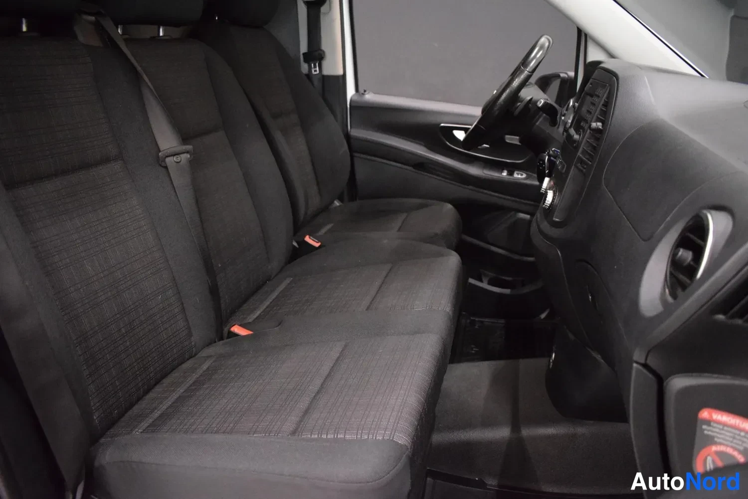 Muu: Sisäkuva etumatkustajan puolelta: tumma kankainen penkkirivi, musta kojelauta ja ohjauspyörä.