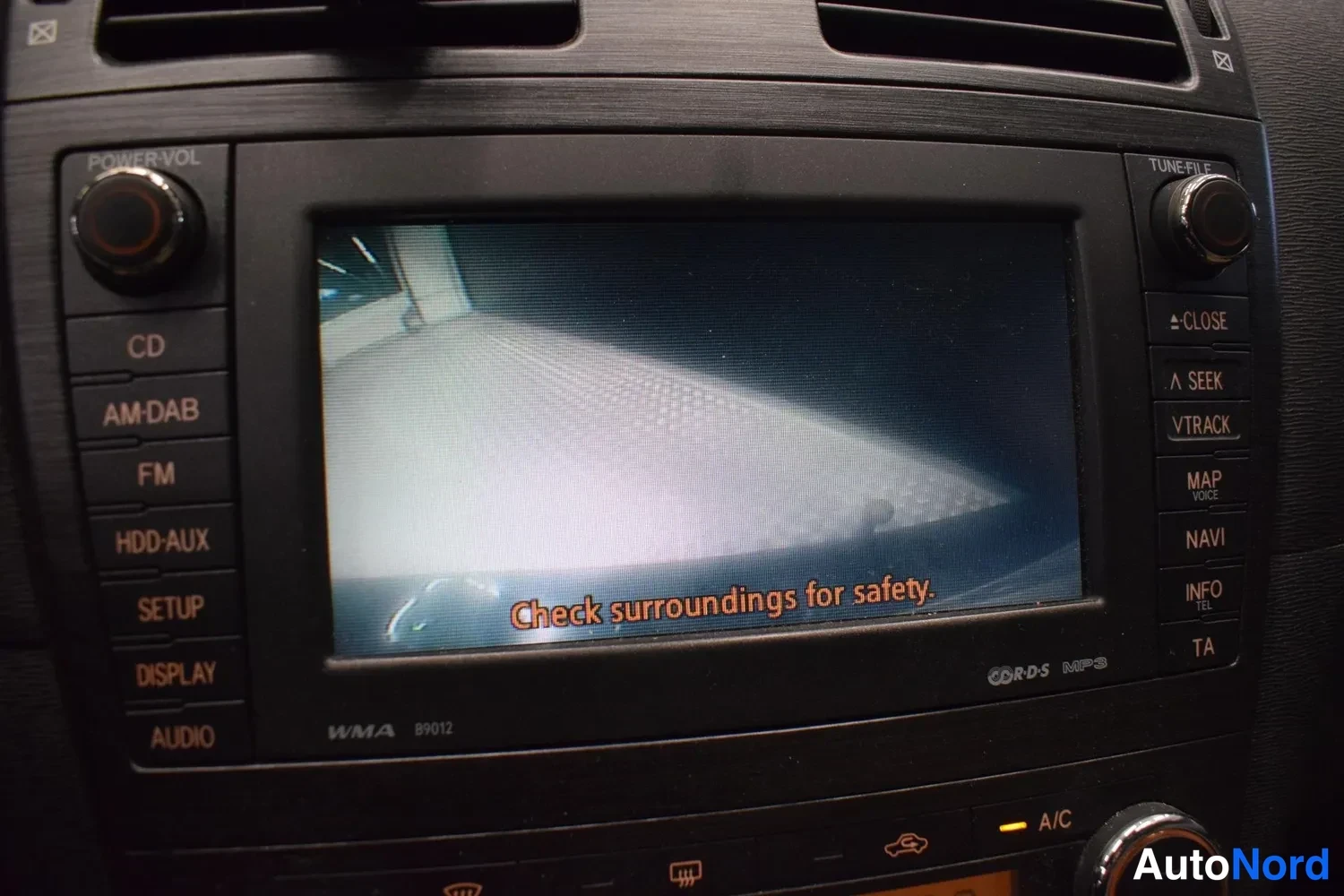 Muu: Kojelaudan infotainment-näyttö näyttää peruutuskameran kuvan tekstillä 'Check surroundings for safety.', ympärillä nappeja kuten CD, FM, NAVI ja A/C.