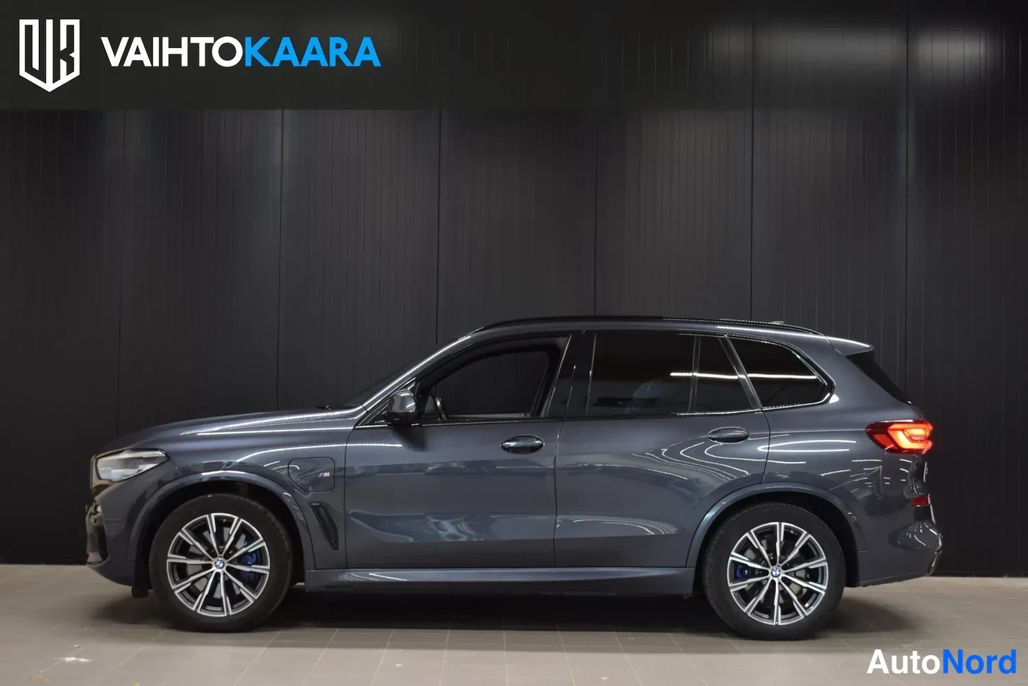 Sivulta: Sinisenharmaa BMW X5 sivukuva vasemmalta sisätiloissa, takavalot syttyneinä.
