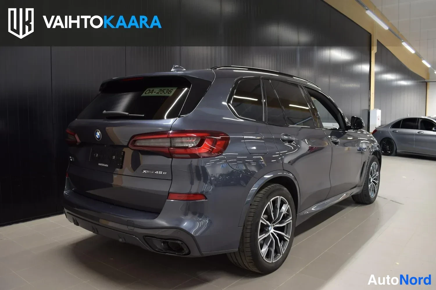 Takaviisto: Sinisenharmaa BMW X5 takanurkkakuva oikealta sisätiloissa, xDrive45e-merkintä peräovessa ja rekisteriotelappu tuulilasissa.